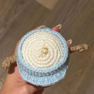 Pu&ograve; includere: Un giocattolo amigurumi all'uncinetto con un corpo bianco e un cappello blu. Il giocattolo ha un anello dorato sulla cima della testa.
