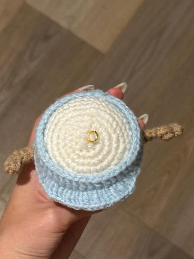 Pu&ograve; includere: Un giocattolo amigurumi all'uncinetto con un corpo bianco e un cappello blu. Il giocattolo ha un anello dorato sulla cima della testa.