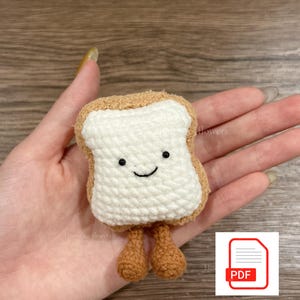 Puede incluir: Una rebanada de pan tostado de ganchillo con una cara sonriente y pequeñas piernas marrones. La tostada tiene un centro blanco y una corteza marrón. La imagen también incluye un icono PDF y las palabras "Digital Download".