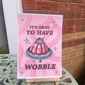 May include: Pink and white print featuring a pink and white jelly dessert with a cherry on top. The print reads "It's okay to have a wobble".