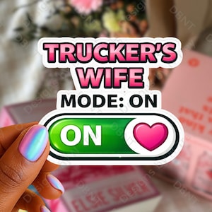 May include: A sticker with the text "TRUCKER'S WIFE MODE: ON" in pink and white. The sticker has a green "ON" button with a pink heart. The sticker is held by a hand with iridescent nail polish.