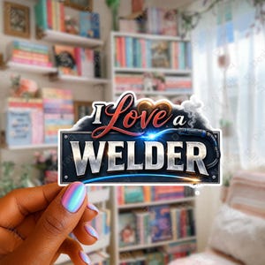 May include: A sticker with the text "I Love a Welder" in red and white. The design includes a welding torch with blue sparks and smoke, set against a dark blue and silver background. The sticker is held in a hand with iridescent nail polish.