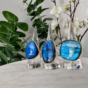 May include: Three silver rings featuring large, polished labradorite gemstones with vibrant blue iridescence. Each ring is displayed on a clear, conical stand. The stones vary in shape, including one rounded, one oval, and one rectangular.