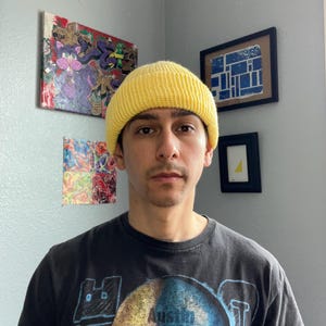 Puede incluir: Un gorro de punto acanalado de color amarillo brillante usado en la cabeza, combinado con una camiseta negra de manga corta con un estampado gr&aacute;fico de una calavera bicolor y el texto Austin 3:16 impreso en una fuente oscura y negrita en el pecho.