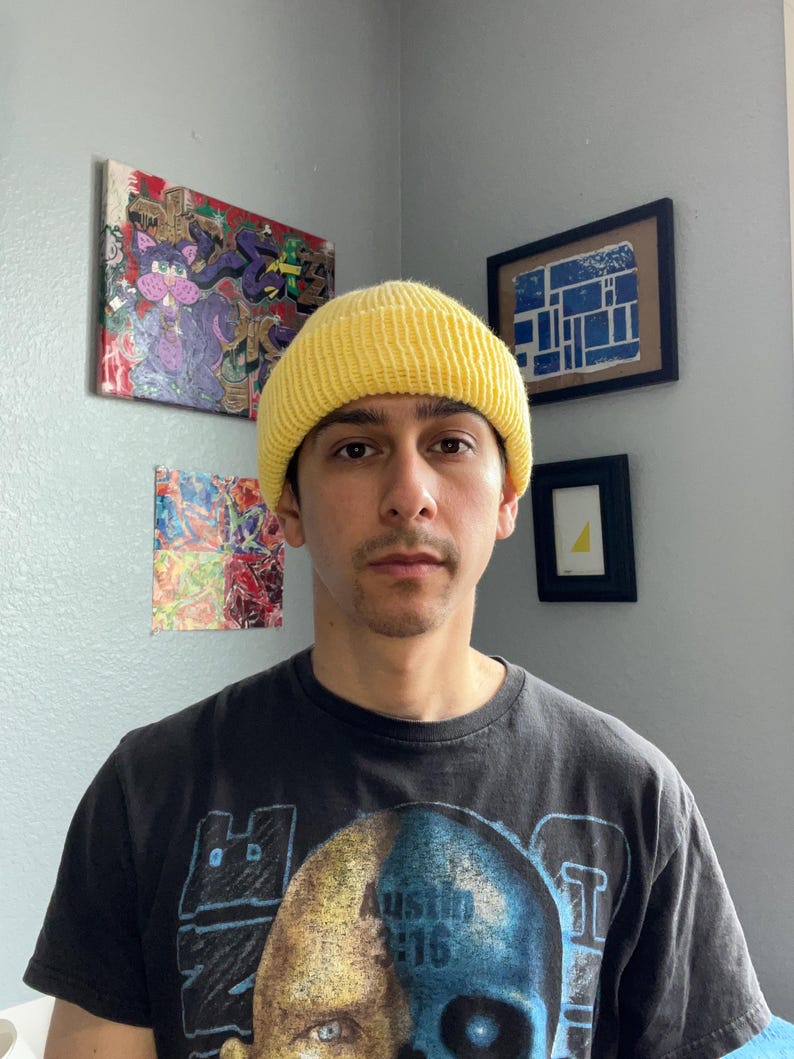 Puede incluir: Un gorro de punto acanalado de color amarillo brillante usado en la cabeza, combinado con una camiseta negra de manga corta con un estampado gr&aacute;fico de una calavera bicolor y el texto Austin 3:16 impreso en una fuente oscura y negrita en el pecho.