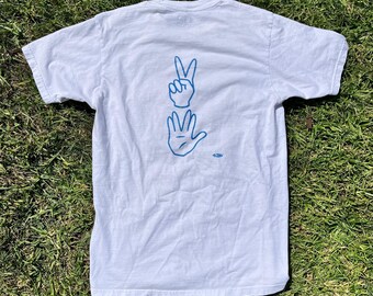 Puede incluir: Camiseta blanca con un contorno azul de dos manos haciendo el signo de la paz y el gesto del número cinco.