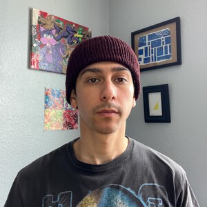 Puede incluir: Un gorro de punto color granate oscuro con textura acanalada. El gorro se lleva puesto en la cabeza y presenta un dise&ntilde;o de borde grueso y doblado que se ajusta c&oacute;modamente sobre las orejas. El tejido tiene un patr&oacute;n de canal&eacute; vertical.