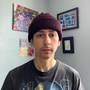 Puede incluir: Un gorro de punto color burdeos oscuro con textura acanalada y borde doblado. El gorro est&aacute; confeccionado con un material de hilo grueso, presentando un patr&oacute;n de puntadas verticales consistente en toda la corona y el borde.