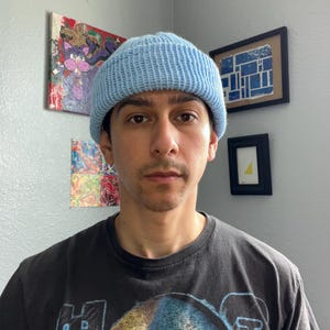 Puede incluir: Un gorro de punto azul claro con textura acanalada y borde doblado. El gorro se lleva puesto en la cabeza, combinado con una camiseta negra de manga corta con un estampado gr&aacute;fico que incluye el texto Austin 3:16 en una fuente gruesa.