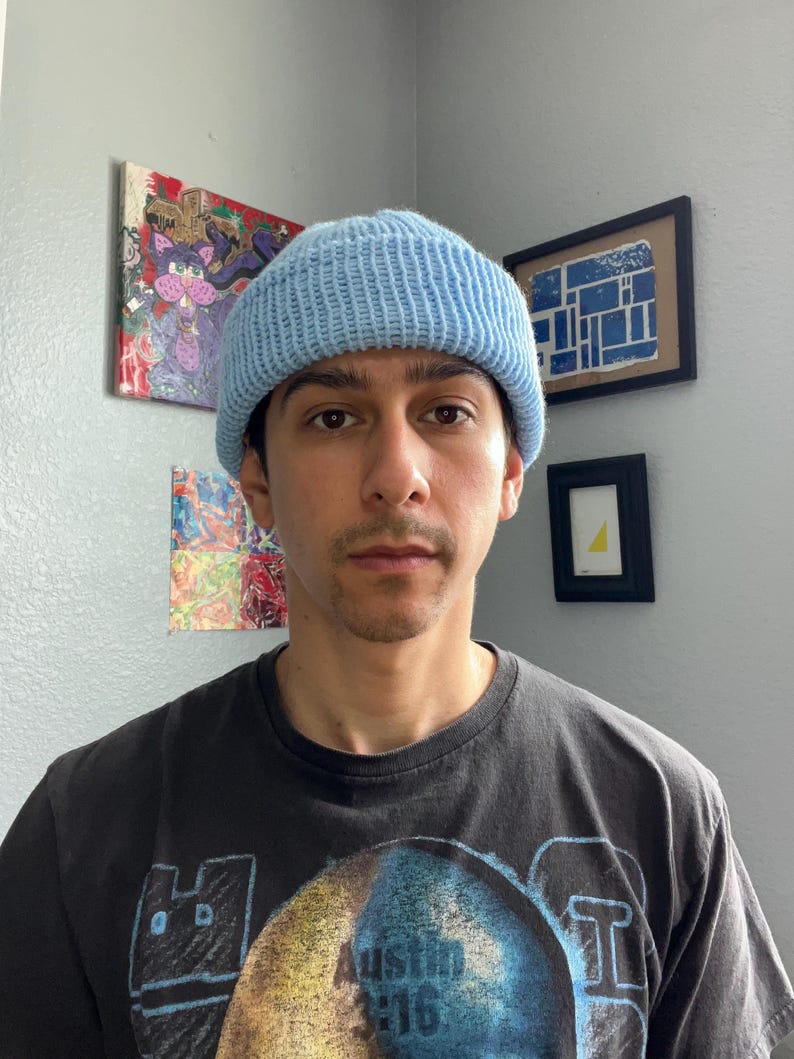 Puede incluir: Un gorro de punto azul claro con textura acanalada y borde doblado. El gorro se lleva puesto en la cabeza, combinado con una camiseta negra de manga corta con un estampado gr&aacute;fico que incluye el texto Austin 3:16 en una fuente gruesa.