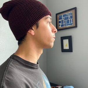 Puede incluir: Un gorro de punto color burdeos oscuro con textura acanalada y ajuste holgado. El gorro est&aacute; confeccionado con un material de hilo grueso y presenta un borde doblado que se ajusta firmemente alrededor de la cabeza, cubriendo la coronilla.