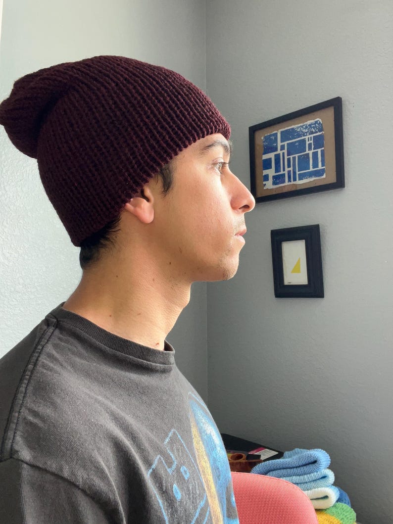 Puede incluir: Un gorro de punto color burdeos oscuro con textura acanalada y ajuste holgado. El gorro est&aacute; confeccionado con un material de hilo grueso y presenta un borde doblado que se ajusta firmemente alrededor de la cabeza, cubriendo la coronilla.
