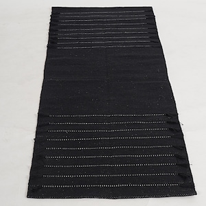 May include: A rectangular black rug with horizontal white dotted lines and fringe on the short ends. The rug is made of a woven material and is displayed on a white surface. This rug is suitable for home decor.