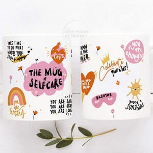 K&ouml;nnte beinhalten: Zwei wei&szlig;e Keramikbecher mit bunten Illustrationen und positiven Affirmationen. Die Becher zeigen S&auml;tze wie "The Mug of Self Care", "You are Worthy", "You are Enough" und "Celebrate Your Wins!"