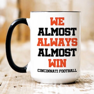 Op de afbeelding: Een witte keramische koffiemok met een zwarte handgreep en een zwarte rand. De mok heeft zwarte tekst die "WE ALMOST ALWAYS ALMOST WIN CINCINNATI FOOTBALL" luidt, met de woorden "ALMOST" en "WIN" in oranje.