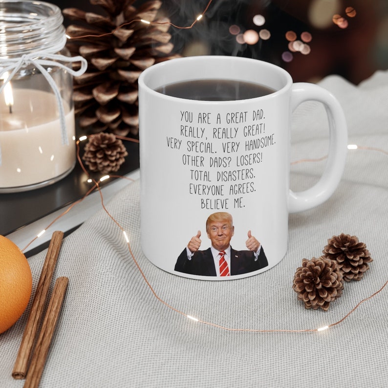 May include: White ceramic coffee mug with a humorous message about dads. The mug features a photo of Donald Trump giving a thumbs up. The text reads: "You are a great dad. Really, really great! Very special. Very handsome. Other dads? Losers! Total disasters. Everyone agrees. Believe me."