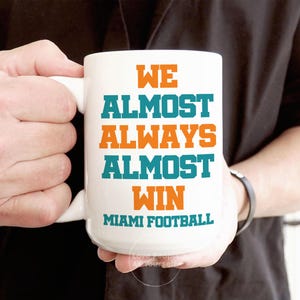 May include: White ceramic coffee mug with teal and orange text that reads "We Almost Always Almost Win Miami Football".