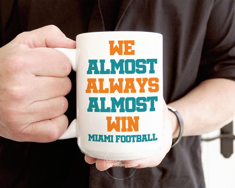 May include: White ceramic coffee mug with teal and orange text that reads "We Almost Always Almost Win Miami Football".