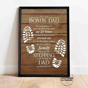 Puede incluir: Una impresi&oacute;n enmarcada con un fondo de madera y texto blanco que dice "To My Bonus Dad". El texto est&aacute; rodeado de huellas blancas. La impresi&oacute;n tiene un marco negro.