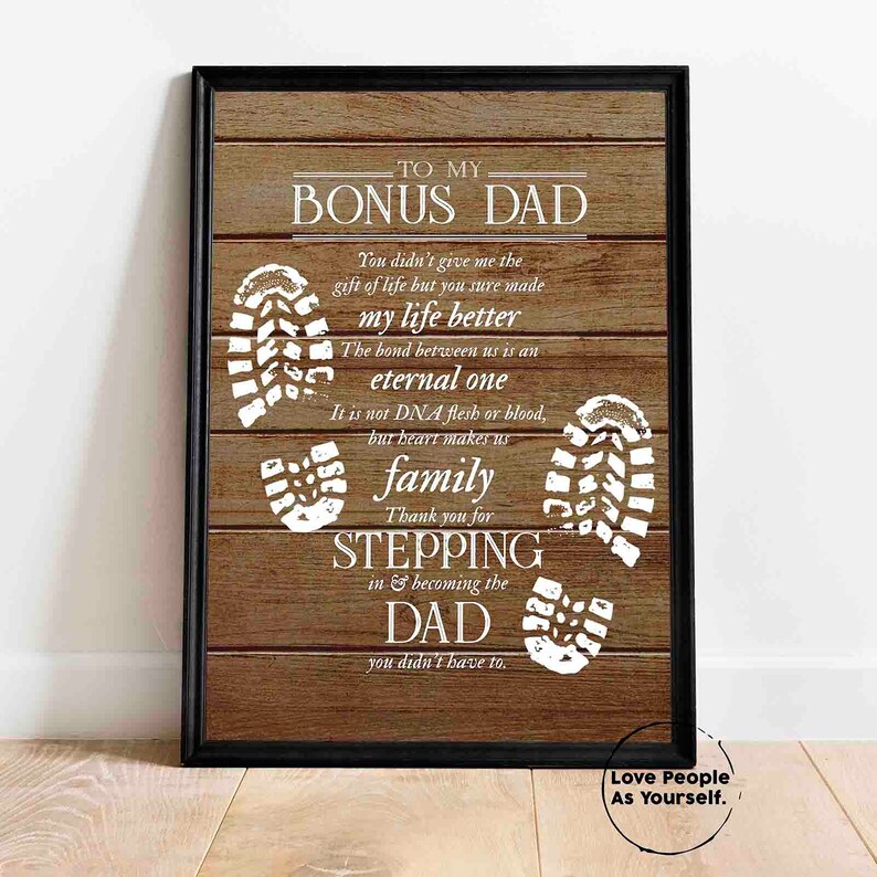 Puede incluir: Una impresi&oacute;n enmarcada con un fondo de madera y texto blanco que dice "To My Bonus Dad". El texto est&aacute; rodeado de huellas blancas. La impresi&oacute;n tiene un marco negro.
