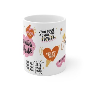 K&ouml;nnte beinhalten: Wei&szlig;e Keramiktasse mit bunten Illustrationen und Text, darunter "Langsam machen & die Blume riechen", "Du bist sicher, du bist okay, du bist geliebt" und "Du bist mutig & tapfer".