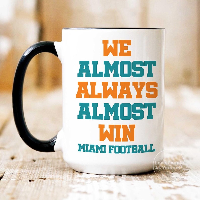 May include: White ceramic coffee mug with a black handle. The mug has teal and orange text that reads "We Almost Always Almost Win Miami Football".