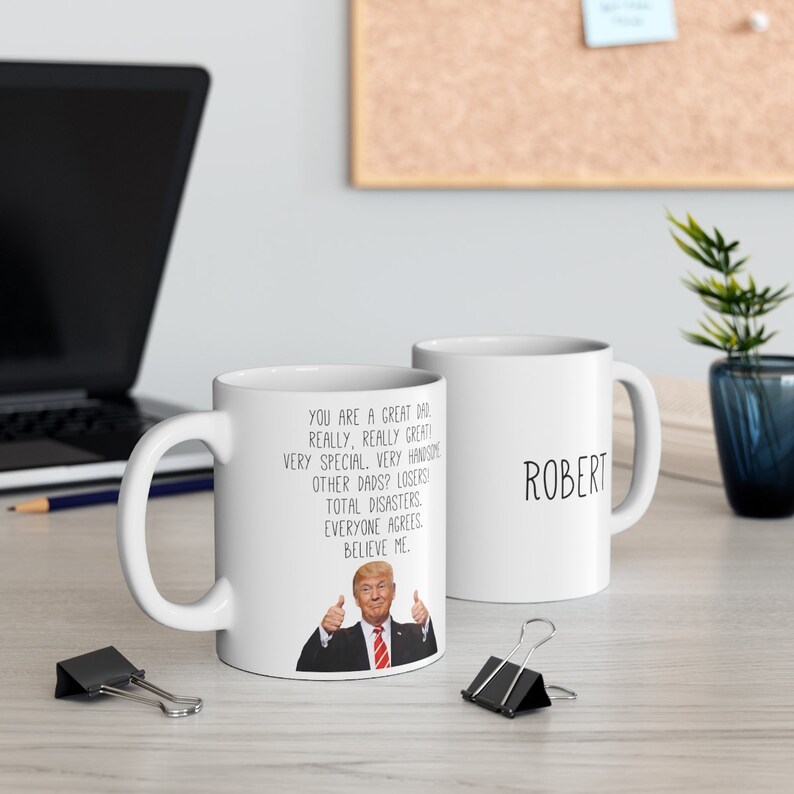 May include: Two white ceramic mugs. One mug features a humorous quote about dads with a photo of a man in a suit giving a thumbs up. The other mug has the name "Robert" written on it.