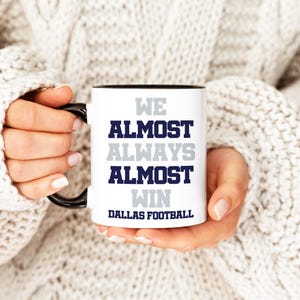 May include: White ceramic mug with a black rim and handle. The mug has the text "We Almost Always Almost Win Dallas Football" printed on it in navy blue and gray.