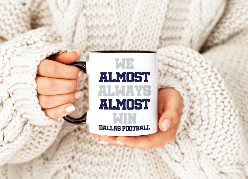 May include: White ceramic mug with a black rim and handle. The mug has the text "We Almost Always Almost Win Dallas Football" printed on it in navy blue and gray.