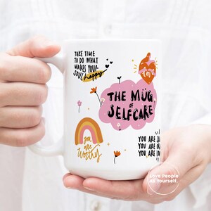 K&ouml;nnte beinhalten: Eine wei&szlig;e Keramiktasse mit einer rosa Wolke und dem Text "The Mug of Selfcare". Die Tasse zeigt au&szlig;erdem einen Regenbogen, Blumen und den Text "Take time to do what makes your soul happy" und "You are worthy".