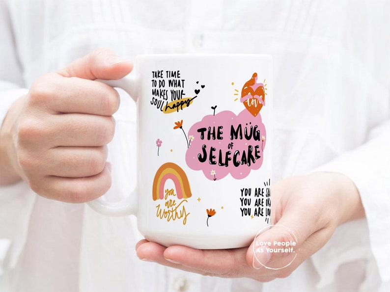 K&ouml;nnte beinhalten: Eine wei&szlig;e Keramiktasse mit einer rosa Wolke und dem Text "The Mug of Selfcare". Die Tasse zeigt au&szlig;erdem einen Regenbogen, Blumen und den Text "Take time to do what makes your soul happy" und "You are worthy".