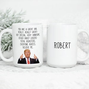 May include: White ceramic coffee mug with a black and white image of a man with a thumbs up gesture. The text on the mug reads "You are a great dad. Really, really great! Very special. Very handsome. Other dads? Losers! Total disasters. Everyone agrees. Believe me." The other side of the mug has the name "Robert" printed on it.