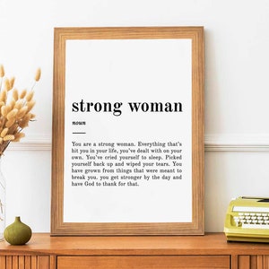 Puede incluir: Una impresi&oacute;n enmarcada con fondo blanco y texto negro. El texto dice "strong woman" y define la palabra como una persona que ha superado desaf&iacute;os y se ha vuelto m&aacute;s fuerte. La impresi&oacute;n est&aacute; en un estante de madera con un jarr&oacute;n de flores secas y una m&aacute;quina de escribir amarilla.