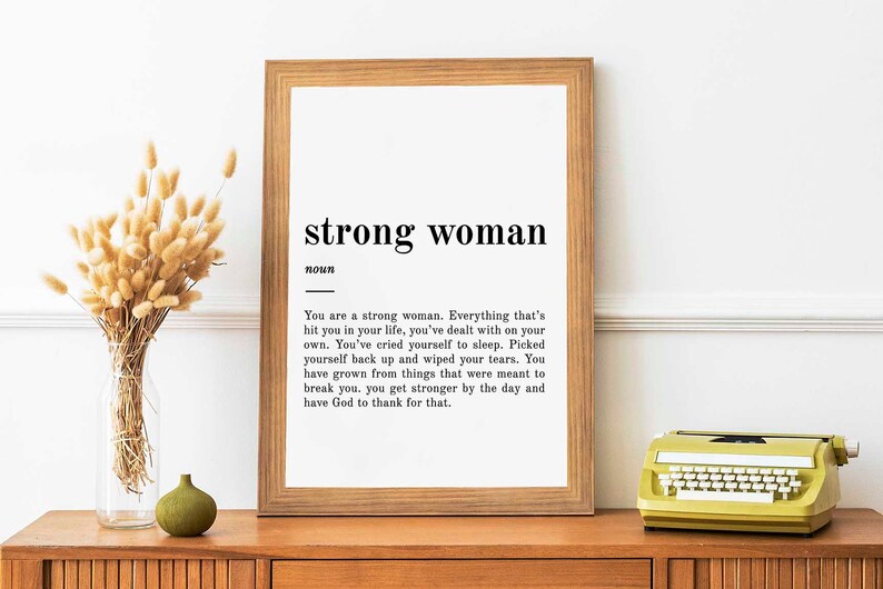 Puede incluir: Una impresi&oacute;n enmarcada con fondo blanco y texto negro. El texto dice "strong woman" y define la palabra como una persona que ha superado desaf&iacute;os y se ha vuelto m&aacute;s fuerte. La impresi&oacute;n est&aacute; en un estante de madera con un jarr&oacute;n de flores secas y una m&aacute;quina de escribir amarilla.