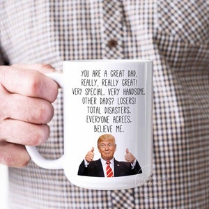 May include: White ceramic coffee mug with a humorous message about dads. The mug features a cartoon image of Donald Trump giving a thumbs up. The text reads: "You are a great dad. Really, really great! Very special. Very handsome. Other dads? Losers! Total disasters. Everyone agrees. Believe me."