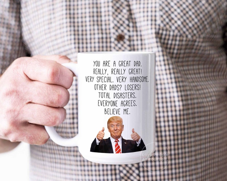 May include: White ceramic coffee mug with a humorous message about dads. The mug features a cartoon image of Donald Trump giving a thumbs up. The text reads: "You are a great dad. Really, really great! Very special. Very handsome. Other dads? Losers! Total disasters. Everyone agrees. Believe me."