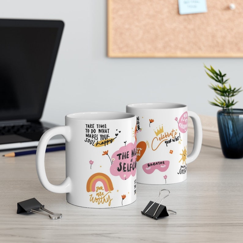 K&ouml;nnte beinhalten: Zwei wei&szlig;e Keramikbecher mit bunten Illustrationen und inspirierenden Zitaten. Ein Becher zeigt einen Regenbogen, eine Wolke mit dem Text "THE MAGIC of SELF-CARE" und den Text "you are worthy". Der andere Becher zeigt eine Sonne, eine Krone und den Text "Celebrate your wins!"