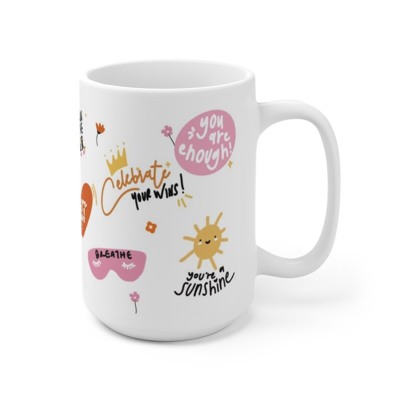 K&ouml;nnte beinhalten: Wei&szlig;e Keramik-Kaffeetasse mit einem farbenfrohen Design mit dem Text "Celebrate your wins!" und anderen positiven Affirmationen. Das Design beinhaltet eine Krone, ein Herz, eine Sonne, ein Augenpaar und Blumen.