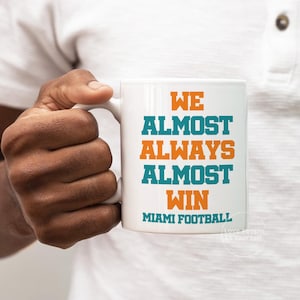 May include: White ceramic coffee mug with the text "We Almost Always Almost Win Miami Football" in orange and teal lettering.
