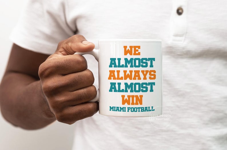 May include: White ceramic coffee mug with the text "We Almost Always Almost Win Miami Football" in orange and teal lettering.