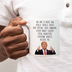 May include: White ceramic coffee mug with a humorous message about dads. The mug features a photo of Donald Trump giving a thumbs up. The text reads: "You are a great dad. Really, really great! Very special. Very handsome. Other dads? Losers! Total disasters. Everyone agrees. Believe me."