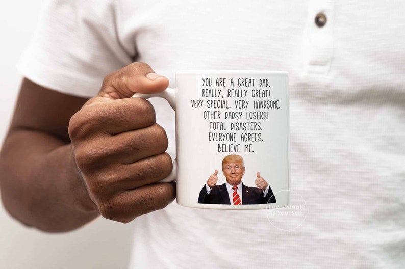 May include: White ceramic coffee mug with a humorous message about dads. The mug features a photo of Donald Trump giving a thumbs up. The text reads: "You are a great dad. Really, really great! Very special. Very handsome. Other dads? Losers! Total disasters. Everyone agrees. Believe me."