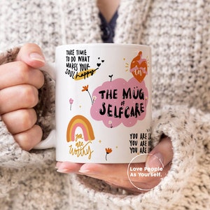 K&ouml;nnte beinhalten: Eine wei&szlig;e Keramiktasse mit einer rosa Wolke und dem Text "The Mug of Selfcare". Die Tasse zeigt auch einen Regenbogen, Blumen und den Text "Take time to do what makes your soul happy", "You are worthy" und "Love People As Yourself."
