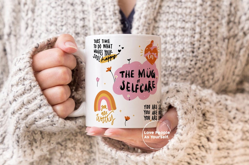 K&ouml;nnte beinhalten: Eine wei&szlig;e Keramiktasse mit einer rosa Wolke und dem Text "The Mug of Selfcare". Die Tasse zeigt auch einen Regenbogen, Blumen und den Text "Take time to do what makes your soul happy", "You are worthy" und "Love People As Yourself."