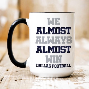 May include: White ceramic coffee mug with a black handle and black rim. The mug has the text "We Almost Always Almost Win Dallas Football" printed on it in gray and blue.