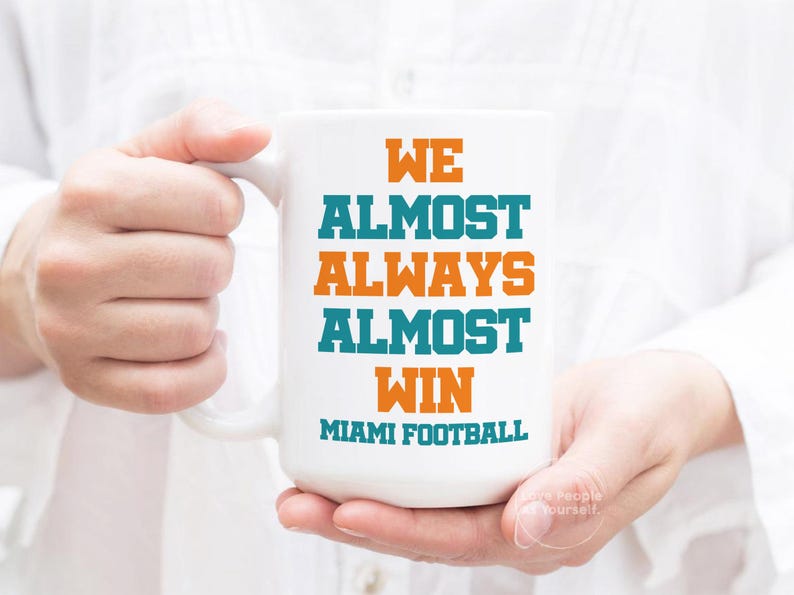May include: White ceramic coffee mug with the text "We Almost Always Almost Win Miami Football" in orange and teal lettering.