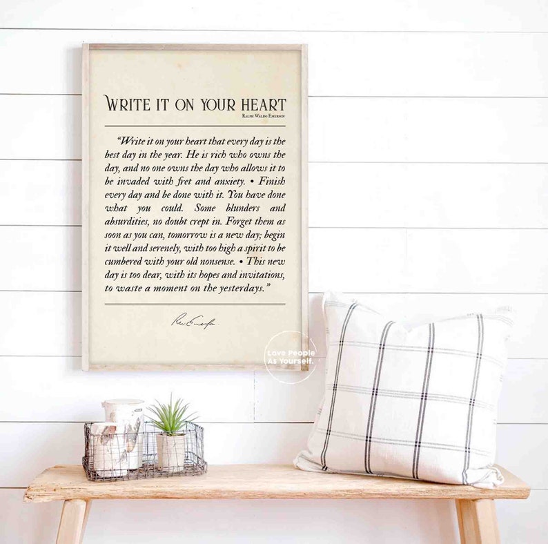 May include: A framed print with a white background and black text. The text reads "Write it on your heart" and includes a quote by Ralph Waldo Emerson. The quote is about living in the present moment and not dwelling on the past. The print is on a wooden shelf with a white and black checkered pillow and a small potted plant.