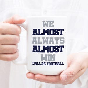 May include: White ceramic mug with the text "We almost always almost win Dallas Football" in navy blue and gray lettering.