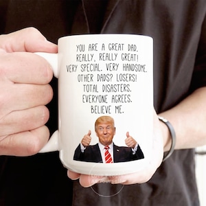 May include: White ceramic coffee mug with a humorous message about dads. The mug features a photo of Donald Trump giving a thumbs up. The text reads: "You are a great dad. Really, really great! Very special. Very handsome. Other dads? Losers! Total disasters. Everyone agrees. Believe me."