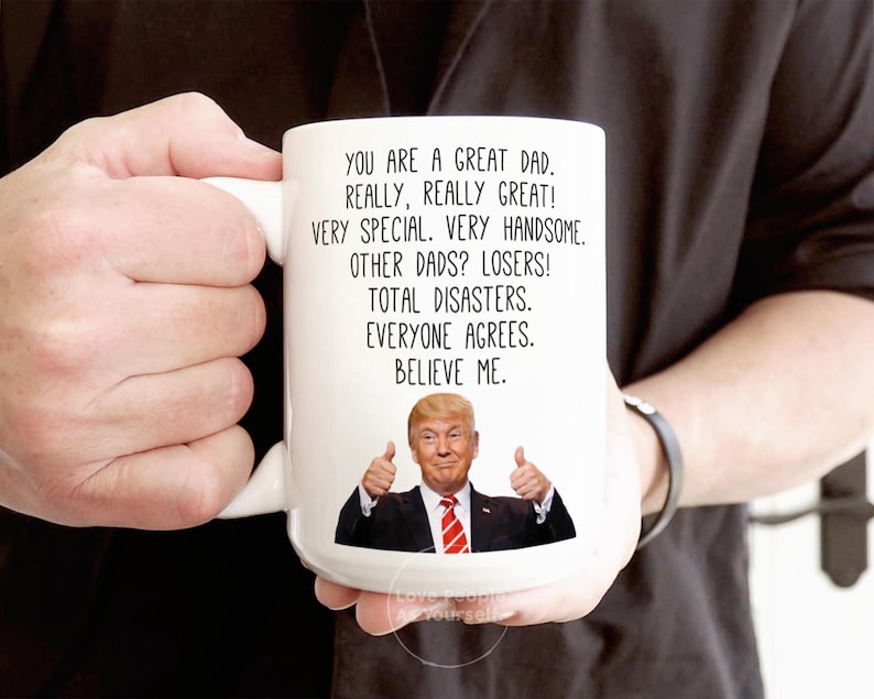 May include: White ceramic coffee mug with a humorous message about dads. The mug features a photo of Donald Trump giving a thumbs up. The text reads: "You are a great dad. Really, really great! Very special. Very handsome. Other dads? Losers! Total disasters. Everyone agrees. Believe me."